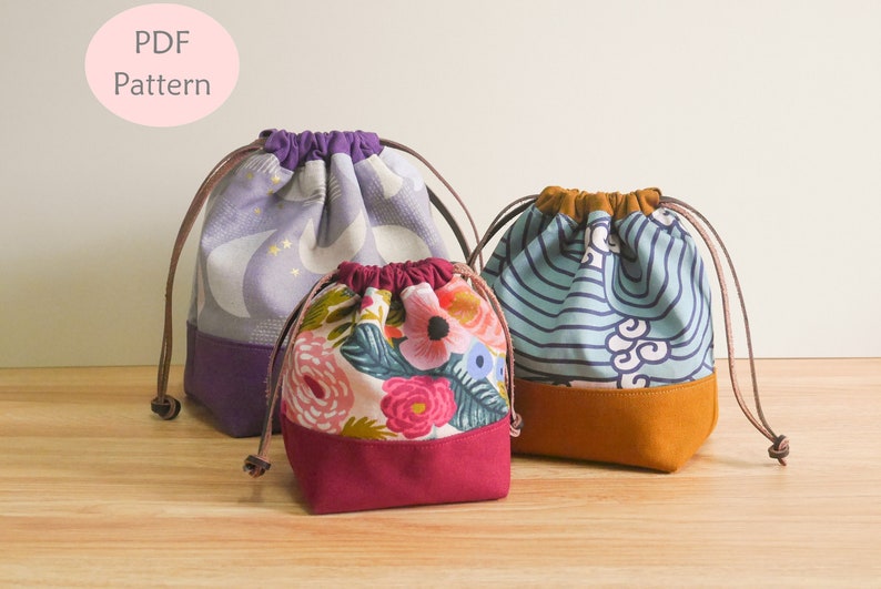 May include: Three drawstring bags made of fabric with different patterns. The largest bag is purple with a white and gold pattern, the middle bag is white with a floral pattern, and the smallest bag is blue with a white and blue wave pattern. All three bags have a solid colored bottom.