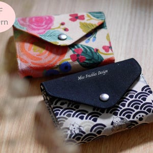 May include: Two small fabric wallets with snap closures. One wallet features a colourful floral pattern with pink, orange, and green tones. The other wallet has a black flap with the text Miss Freckles Design and a white pattern of waves and cats.