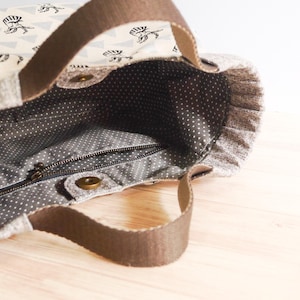 May include: A fabric tote bag with brown handles and a patterned exterior. The bag's exterior features a repeating pattern of black illustrations on a cream background. The interior is gray with white polka dots, and a zipper closure is visible.