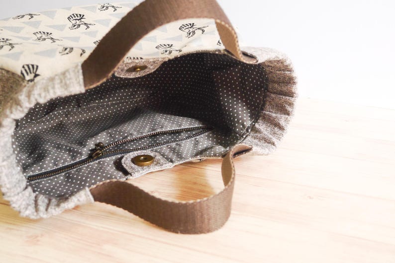 May include: A fabric tote bag with brown handles and a patterned exterior. The bag's exterior features a repeating pattern of black illustrations on a cream background. The interior is gray with white polka dots, and a zipper closure is visible.