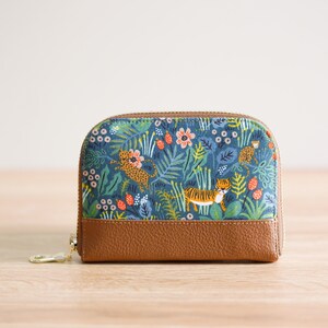 May include: A small, blue and brown zippered pouch with a floral and animal print. The print features tigers, leopards, and flowers on a blue background. The pouch has a brown leather bottom.