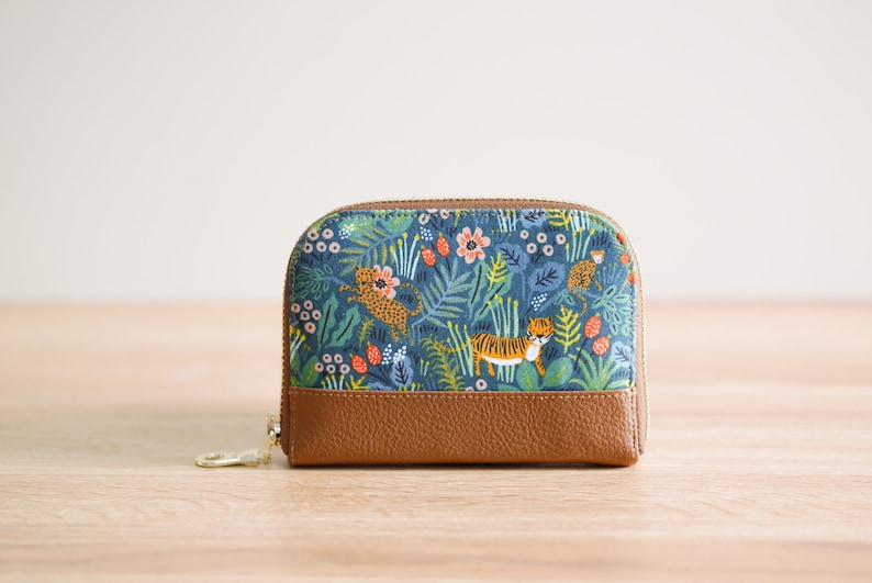 May include: A small, blue and brown zippered pouch with a floral and animal print. The print features tigers, leopards, and flowers on a blue background. The pouch has a brown leather bottom.