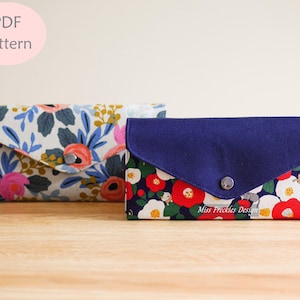 May include: Two rectangular fabric wallets with envelope-style flaps and snap closures. One wallet features a floral pattern with pink, blue, and green on white. The other has a dark blue flap over a floral print with red, white, and small cat figures.