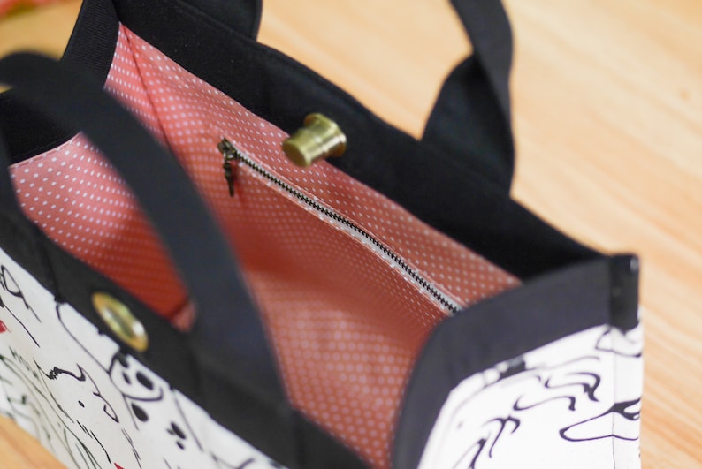 Pu&ograve; includere: Una borsa tote bianca e nera con una fodera a pois rosa. La borsa ha una tasca con cerniera e un manico superiore.