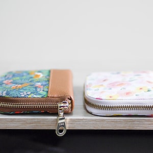 May include: Two zippered wallets, one with a floral pattern and a brown leather accent, and the other with a white background and a floral pattern in shades of pink, yellow, and green.