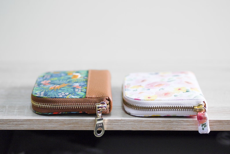 May include: Two zippered wallets, one with a floral pattern and a brown leather accent, and the other with a white background and a floral pattern in shades of pink, yellow, and green.