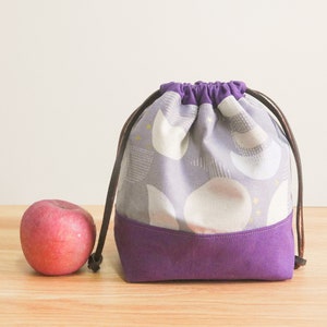 May include: A purple drawstring bag with a white and gray geometric pattern. The bag has brown drawstrings and a purple bottom.