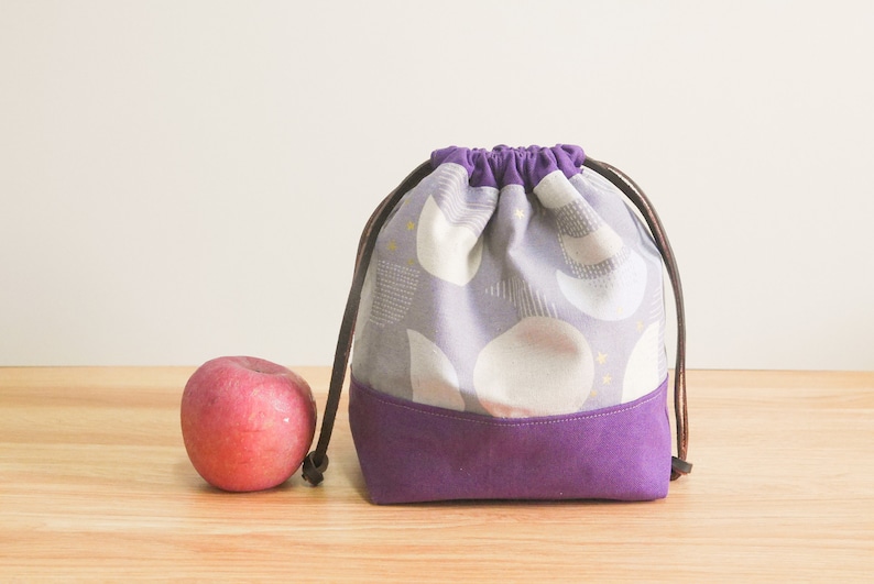 May include: A purple drawstring bag with a white and gray geometric pattern. The bag has brown drawstrings and a purple bottom.