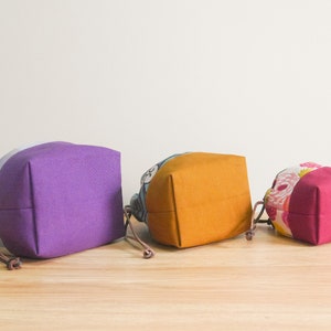 May include: Three fabric drawstring bags in different sizes and colors. The largest bag is purple, the middle bag is yellow, and the smallest bag is pink.