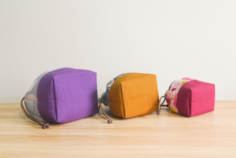 May include: Three fabric drawstring bags in different sizes and colors. The largest bag is purple, the middle bag is yellow, and the smallest bag is pink.