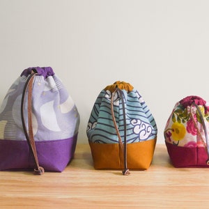 May include: Three drawstring bags in different patterns and colors. The bag on the left has a purple and white geometric pattern, the middle bag has a blue and white striped pattern, and the bag on the right has a floral pattern with pink, red, and yellow flowers. All three bags have brown leather drawstrings.