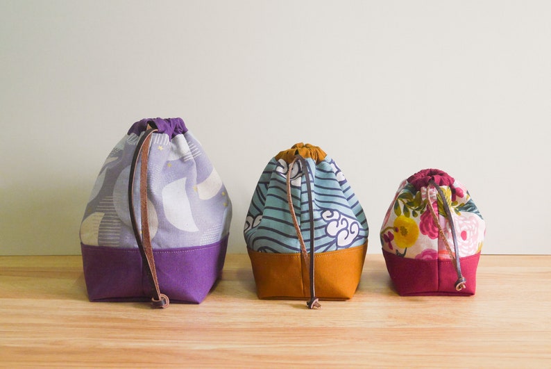 May include: Three drawstring bags in different patterns and colors. The bag on the left has a purple and white geometric pattern, the middle bag has a blue and white striped pattern, and the bag on the right has a floral pattern with pink, red, and yellow flowers. All three bags have brown leather drawstrings.