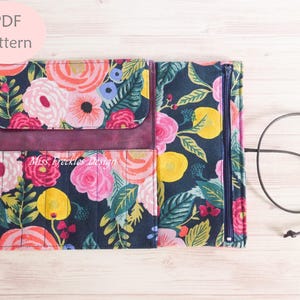 May include: A fabric roll-up organizer featuring a dark navy background with a vibrant floral print in shades of pink, yellow, and green. It includes a flap, a zippered compartment, and a long dark cord for closure. The text Miss Freckles Design is visible.