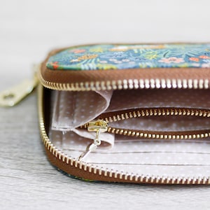 May include: A small, floral patterned zippered wallet with a brown leather trim. The wallet is open, revealing a white fabric interior with two zippered compartments.
