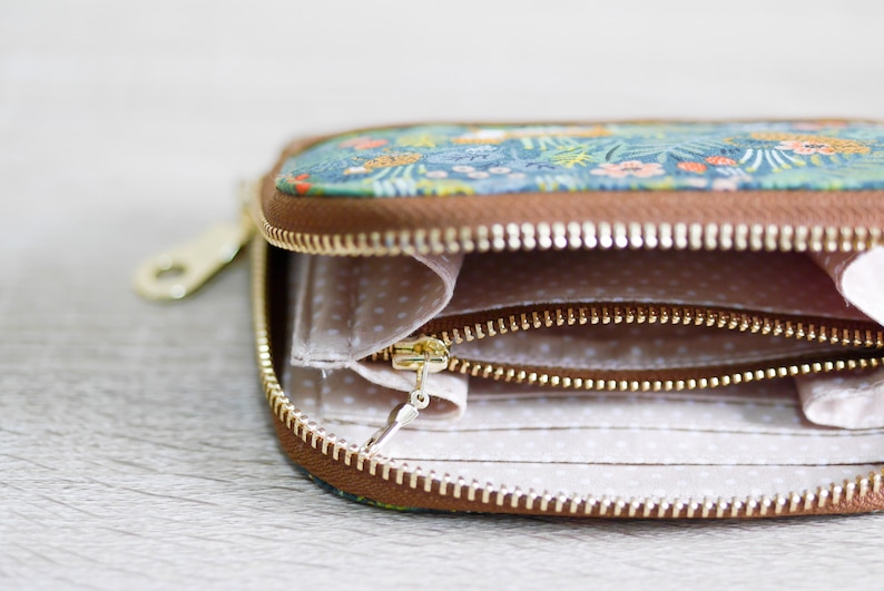 May include: A small, floral patterned zippered wallet with a brown leather trim. The wallet is open, revealing a white fabric interior with two zippered compartments.
