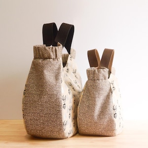 May include: Three fabric bags with brown handles. The bags are different sizes and have a textured, light brown fabric on the front and back. The middle bag has a cream-colored fabric with a black pattern.