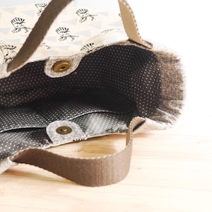 May include: Open tote bag with a light beige exterior patterned with black figures and triangles. The interior is gray with white polka dots and has pockets. Brown handles and a magnetic snap closure are visible.