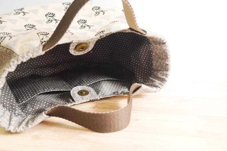 May include: Open tote bag with a light beige exterior patterned with black figures and triangles. The interior is gray with white polka dots and has pockets. Brown handles and a magnetic snap closure are visible.