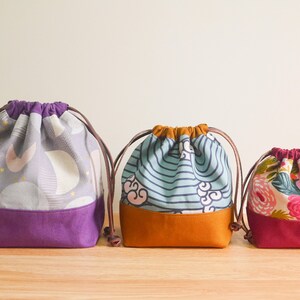 May include: Three drawstring bags with different patterns. The bag on the left has a purple and gray pattern with gold stars. The middle bag has a blue and white striped pattern with a brown bottom. The bag on the right has a pink and white floral pattern with a red bottom.