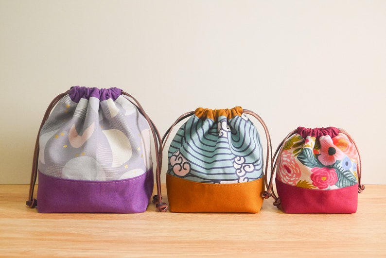 May include: Three drawstring bags with different patterns. The bag on the left has a purple and gray pattern with gold stars. The middle bag has a blue and white striped pattern with a brown bottom. The bag on the right has a pink and white floral pattern with a red bottom.
