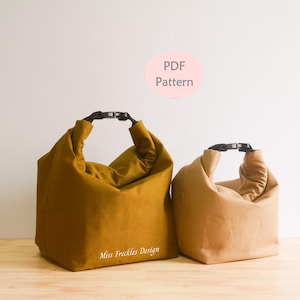 Pode incluir: Duas bolsas com fecho de enrolar e fivelas de pl&aacute;stico preto. A bolsa maior &eacute; amarelo-mostarda com o texto Miss Freckles Design, e a bolsa menor &eacute; bege. Um c&iacute;rculo rosa acima cont&eacute;m o texto PDF Pattern.