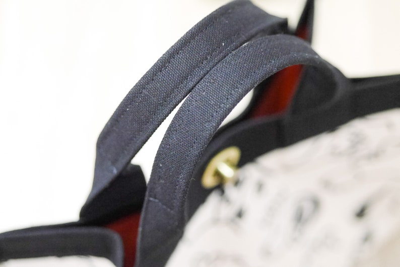 Pu&ograve; includere: Primo piano delle maniglie in tela nera di una borsa tote. Le maniglie sono cucite a doppio punto e hanno una fodera rossa.