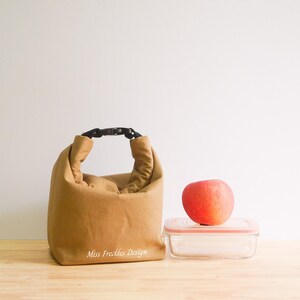 Pode incluir: Uma lancheira de tecido castanho-claro com uma al&ccedil;a de fivela preta e o texto Miss Freckles Design impresso em branco na frente. Ao lado da bolsa, h&aacute; um recipiente de vidro transparente com tampa rosa, sobre o qual repousa uma ma&ccedil;&atilde; vermelha.