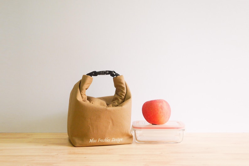 Pode incluir: Uma lancheira de tecido castanho-claro com uma al&ccedil;a de fivela preta e o texto Miss Freckles Design impresso em branco na frente. Ao lado da bolsa, h&aacute; um recipiente de vidro transparente com tampa rosa, sobre o qual repousa uma ma&ccedil;&atilde; vermelha.
