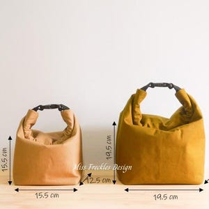 Pode incluir: Duas lancheiras com fecho de enrolar e fivelas de pl&aacute;stico preto. A bolsa menor &eacute; bege e mede 15,5 cm por 15,5 cm. A bolsa maior &eacute; amarelo-mostarda e mede 19,5 cm por 19,5 cm. O texto Miss Freckles Design est&aacute; impresso entre as duas bolsas.