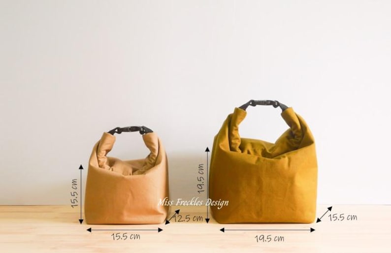 Pode incluir: Duas lancheiras com fecho de enrolar e fivelas de pl&aacute;stico preto. A bolsa menor &eacute; bege e mede 15,5 cm por 15,5 cm. A bolsa maior &eacute; amarelo-mostarda e mede 19,5 cm por 19,5 cm. O texto Miss Freckles Design est&aacute; impresso entre as duas bolsas.