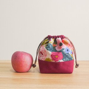 May include: A small burgundy drawstring bag with a floral print. The bag is made of fabric and has a drawstring closure. The bag is sitting on a wooden surface next to a red apple.