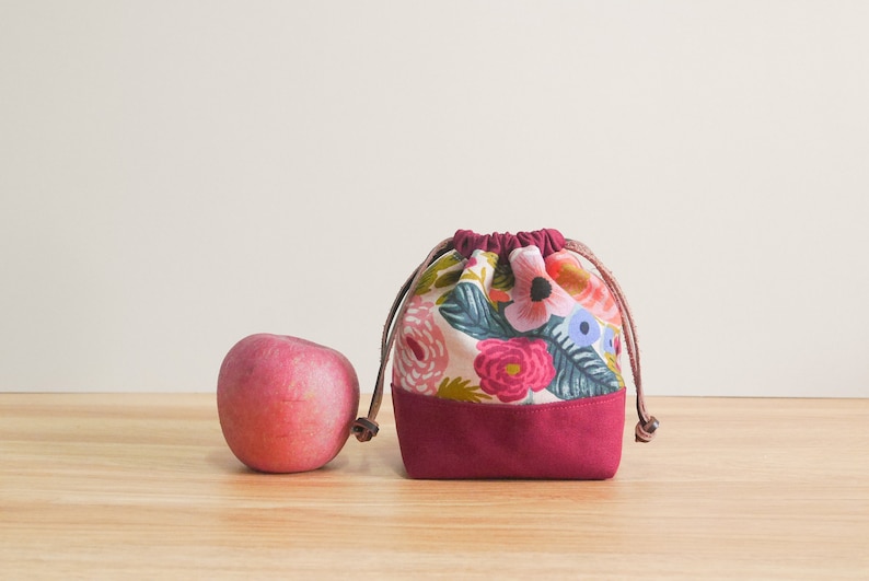May include: A small burgundy drawstring bag with a floral print. The bag is made of fabric and has a drawstring closure. The bag is sitting on a wooden surface next to a red apple.