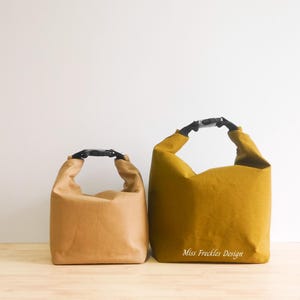 Pode incluir: Duas bolsas com fecho de enrolar e fivelas de pl&aacute;stico preto. A bolsa menor &eacute; castanho-claro e a maior &eacute; amarelo-mostarda com o texto branco Miss Freckles Design impresso na frente. Ambas as bolsas apresentam um design de base retangular.