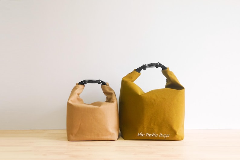 Pode incluir: Duas bolsas com fecho de enrolar e fivelas de pl&aacute;stico preto. A bolsa menor &eacute; castanho-claro e a maior &eacute; amarelo-mostarda com o texto branco Miss Freckles Design impresso na frente. Ambas as bolsas apresentam um design de base retangular.