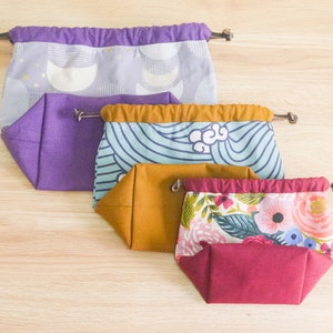 May include: Four fabric drawstring bags in different sizes and patterns. The largest bag is purple with a white and gold moon pattern. The next bag is teal with a white wave pattern. The third bag is mustard yellow with a white floral pattern. The smallest bag is burgundy with a white floral pattern.