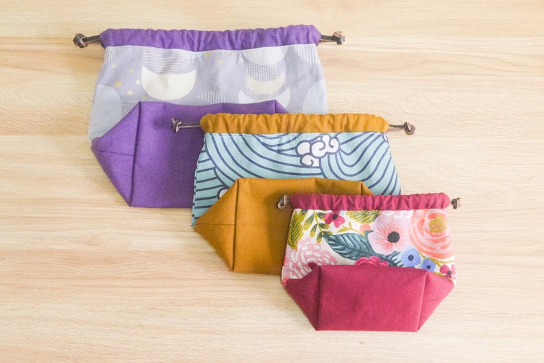 May include: Four fabric drawstring bags in different sizes and patterns. The largest bag is purple with a white and gold moon pattern. The next bag is teal with a white wave pattern. The third bag is mustard yellow with a white floral pattern. The smallest bag is burgundy with a white floral pattern.
