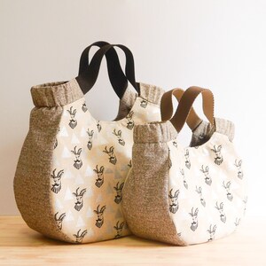 May include: Two tote bags of different sizes. The bags have a beige and brown color scheme. The main body of the bags is beige with a repeating black goat illustration. The sides and top trim are brown. The handles are brown and black.