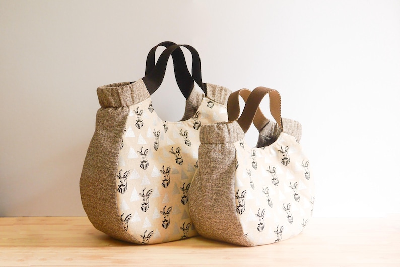 May include: Two tote bags of different sizes. The bags have a beige and brown color scheme. The main body of the bags is beige with a repeating black goat illustration. The sides and top trim are brown. The handles are brown and black.