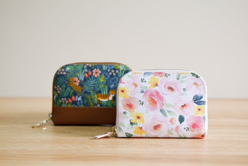 May include: Two small zippered wallets with floral patterns. One wallet has a blue and green floral pattern with tigers, the other has a white background with pink and yellow flowers.