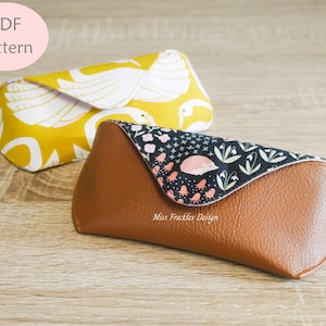 Può includere: Due custodie per occhiali. Una è gialla con un motivo a cigni bianchi. L'altra è in similpelle marrone con una patta nera caratterizzata da un riccio rosa e una stampa floreale. Il testo Miss Frackles Design è stampato sulla custodia marrone. Un cerchio rosa riporta PDF Pattern.