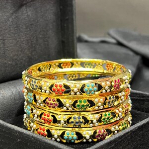 May include: A set of four gold bangles with black enamel and colorful gemstones. The bangles are decorated with a floral design and white pearls.
