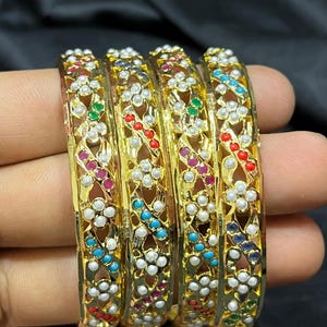 May include: Set of four gold bangles with intricate designs. Each bangle features a row of white pearls and colorful enamel accents.