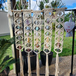 May include: Five vertical macrame wall hangings made of off-white cotton cord, each featuring a series of raw, multi-coloured crystals secured within diamond-shaped knots. Each piece terminates in a long, loose fringe at the bottom.