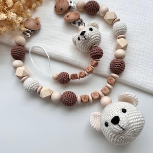 Könnte beinhalten: Ein Baby-Schnullerhalter mit einem weißen, gehäkelten Teddybärenkopf, Holzperlen und braunen, gehäkelten Perlen. Der Name "ARIA" ist in Holzbuchstabenperlen geschrieben. Der Clip besteht aus Holz und Metall.