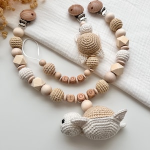 May include: Two baby pacifier clips with wooden beads, geometric shapes, and crocheted turtle accents. One clip features the name "LOU" spelled out in wooden letter beads. The clips have wooden closures and are designed for infants.