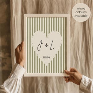 May include: A framed vertical print featuring a sage green and white vertical striped background. In the centre, a large cream-coloured heart with a dotted border contains the black text J & L and the date 23.10.2014 in a simple font.