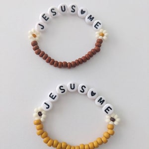 May include: Two beaded bracelets with the words "JESUS loves ME" and white daisy beads. The top bracelet has brown beads and the bottom bracelet has yellow beads.