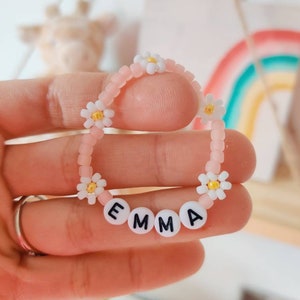 May include: Pink beaded bracelet with white daisy beads and white letter beads spelling the name "EMMA".