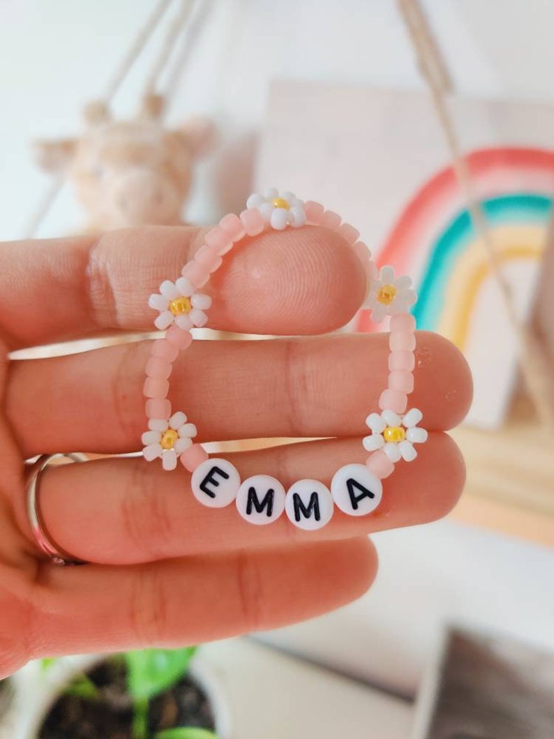 May include: Pink beaded bracelet with white daisy beads and white letter beads spelling the name "EMMA".