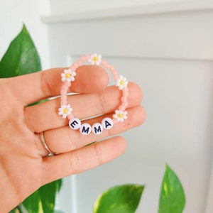 May include: Pink beaded bracelet with white daisy beads and white letter beads spelling the name "EMMA".